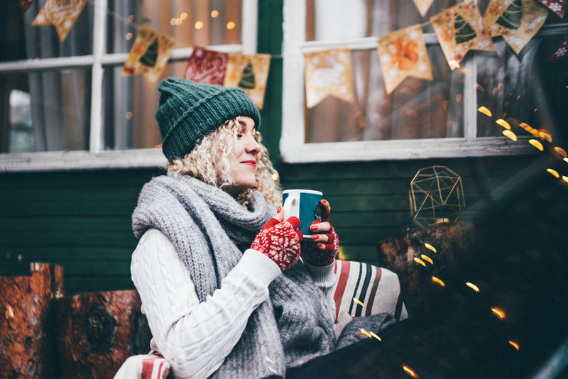 Smiling woman dressed warmly in a green knit hat and scarf, enjoying a hot drink in a cozy outdoor winter retreat adorned with festive decorations.