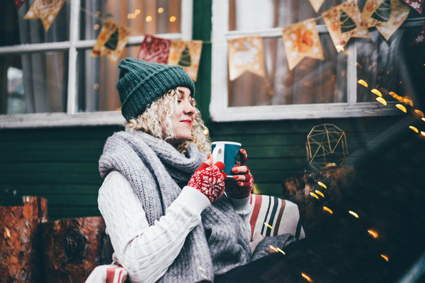 Smiling woman dressed warmly in a green knit hat and scarf, enjoying a hot drink in a cozy outdoor winter retreat adorned with festive decorations.